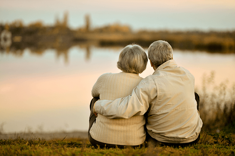 An older couple sitting on the grass looking at a lake.